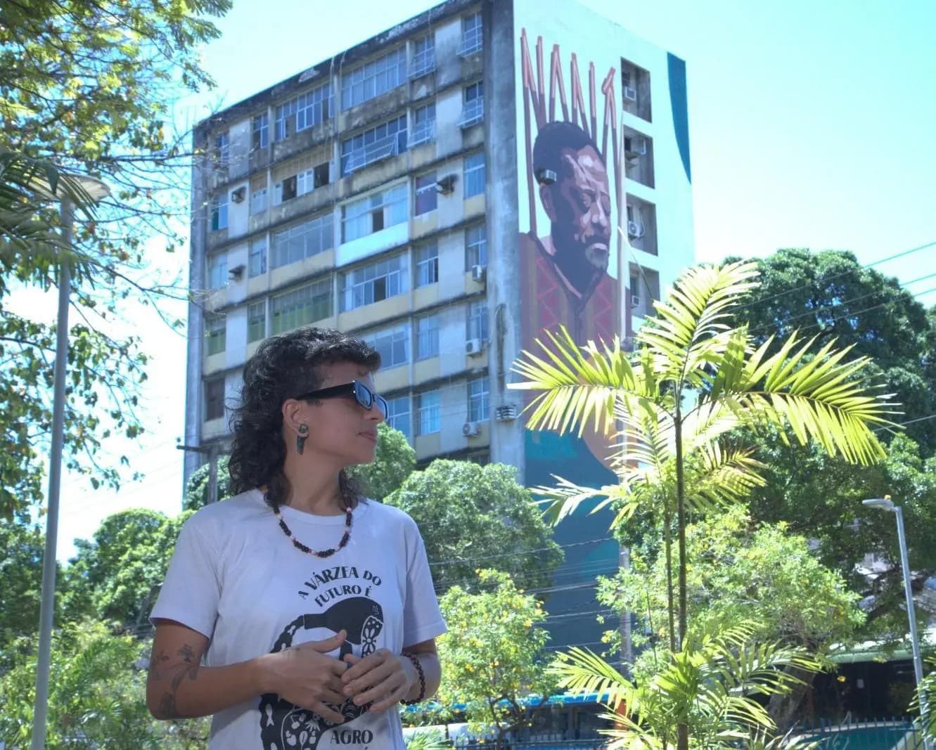 Foto da artista Micaela Almeida usando óculos escuros no centro do recife com sua obra de Mega Mural de Naná Vasconcelos ao fundo